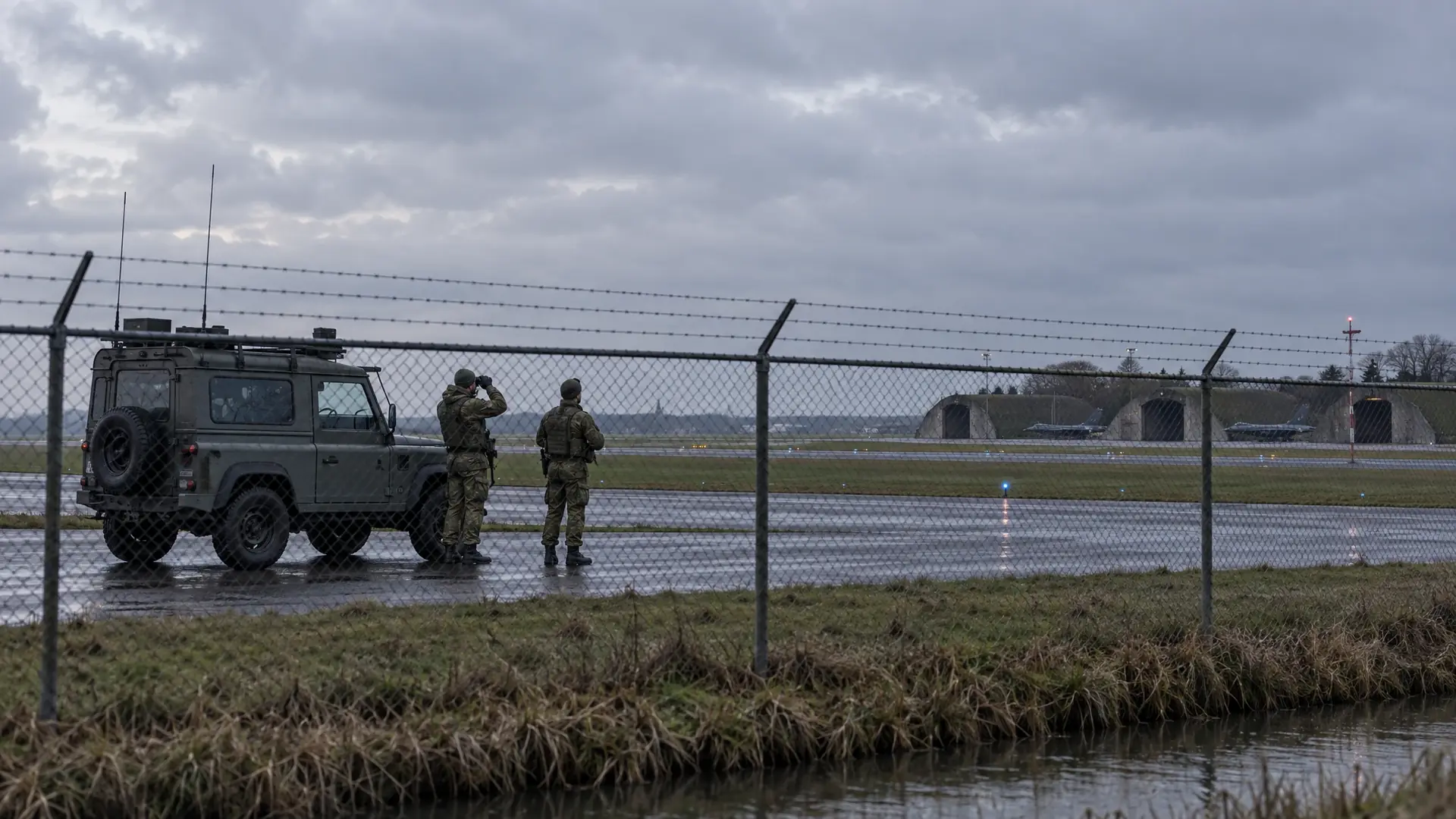 Two soldiers scanning an empty sky from a concrete apron beside a chain-link fence, with a patrol vehicle and the hangars of a Dutch military airbase behind them